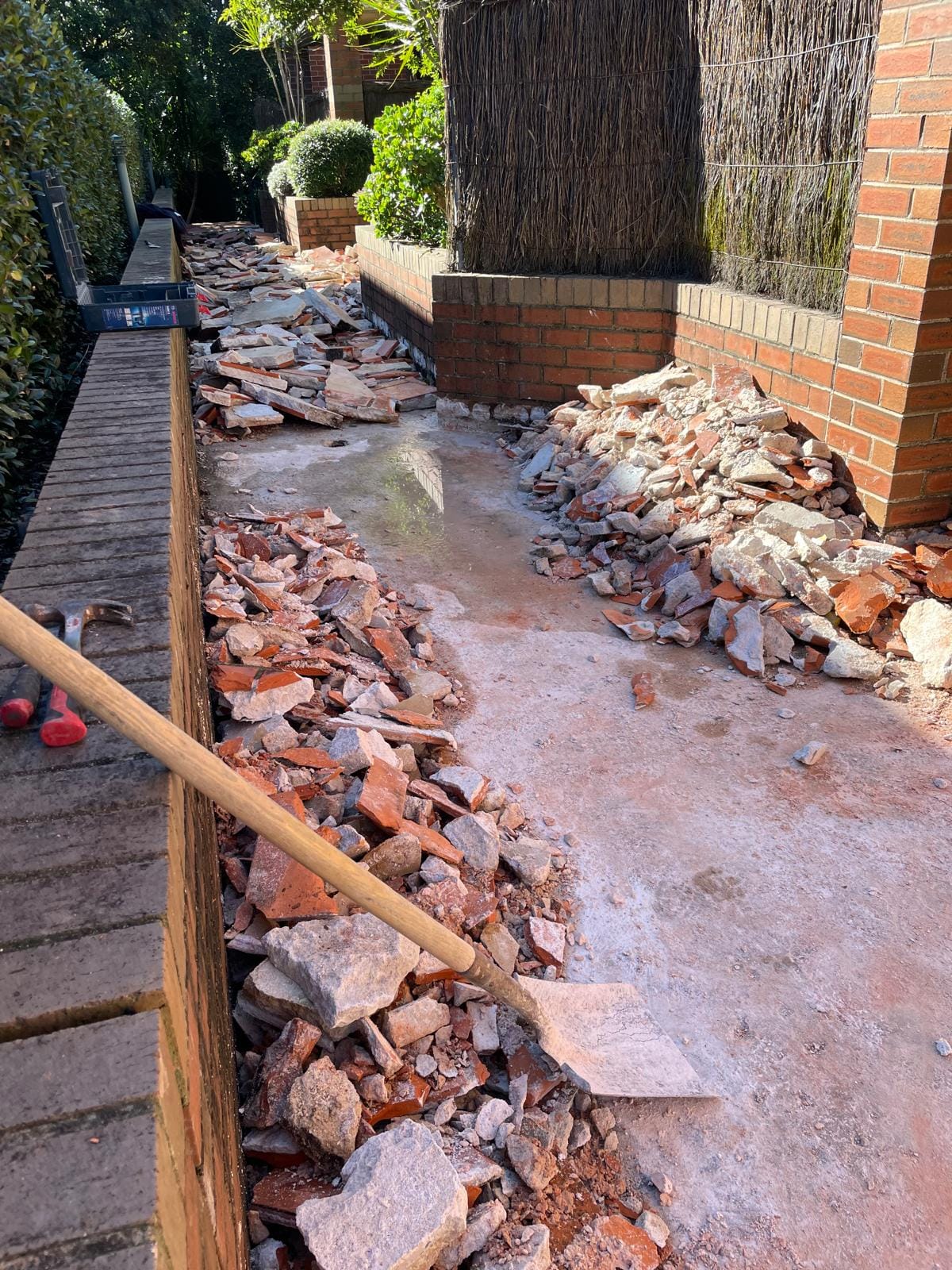 Mid-demolition: broken terracotta and bedding mortar heaped along both sides of the path, shovel and hammer at hand, the slab appearing underneath.