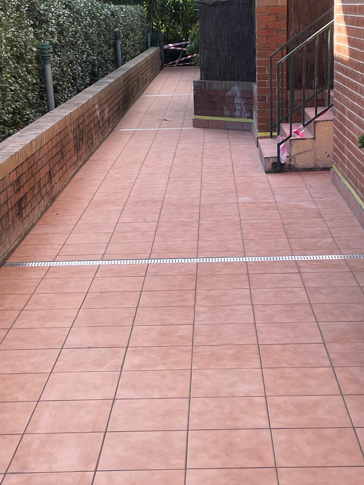 A stainless strip drain set flush across the finished walkway, tiles cut to meet it square on both sides and carried neatly around the entry steps.