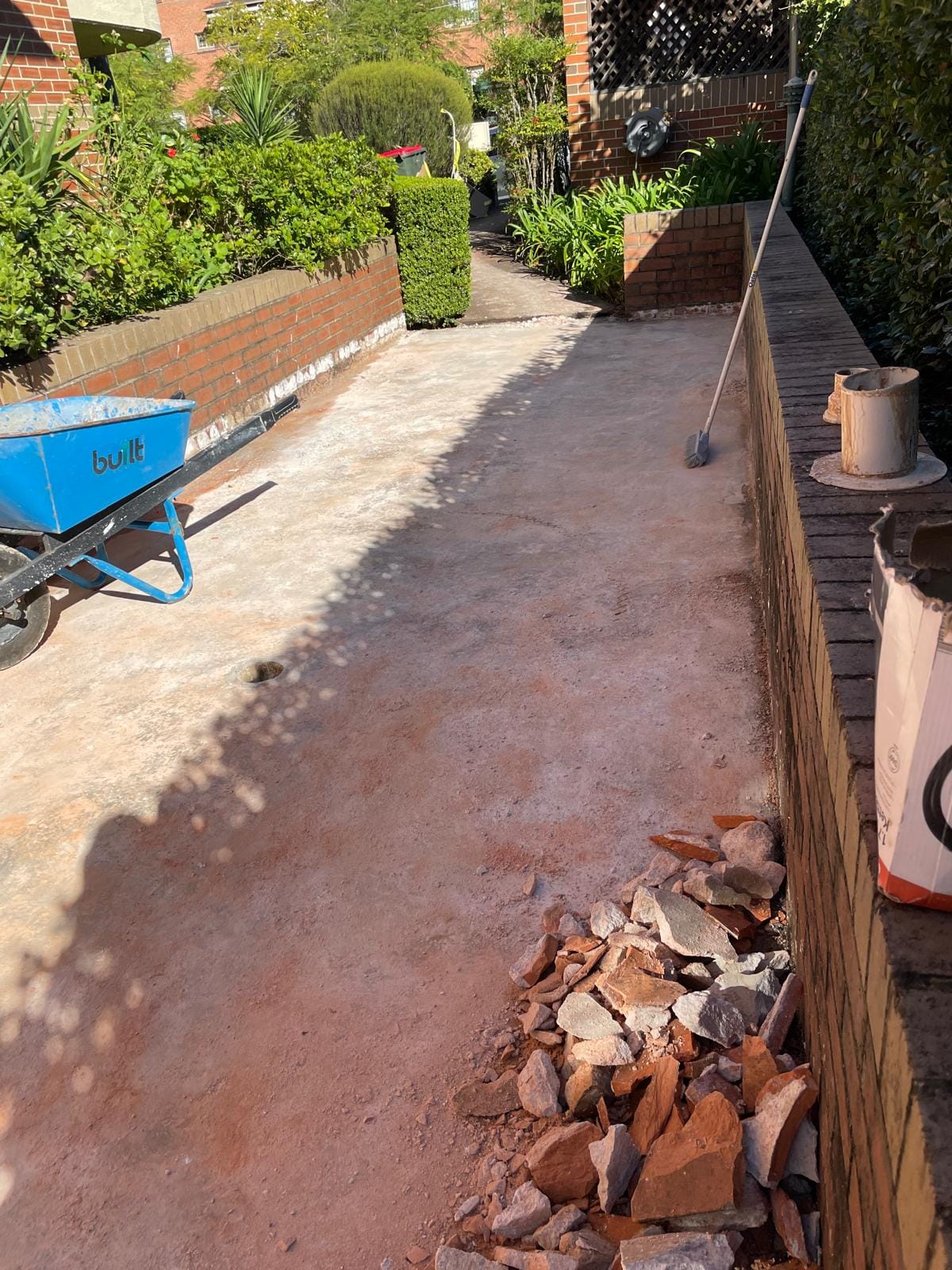 The old tiles lifted and stacked for removal beside the exposed concrete, wheelbarrow and broom on site, the garden either side left undisturbed.