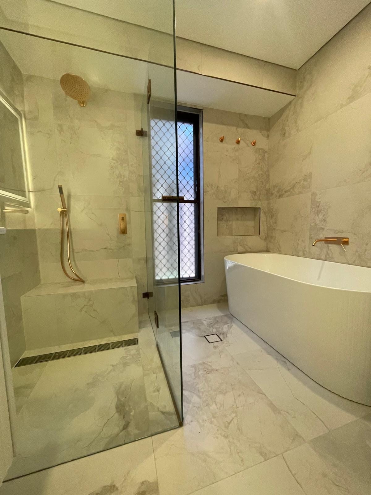 Frameless shower with a full-width stone bench, brass rail shower and a linear drain, alongside a freestanding bath with a recessed niche above it.