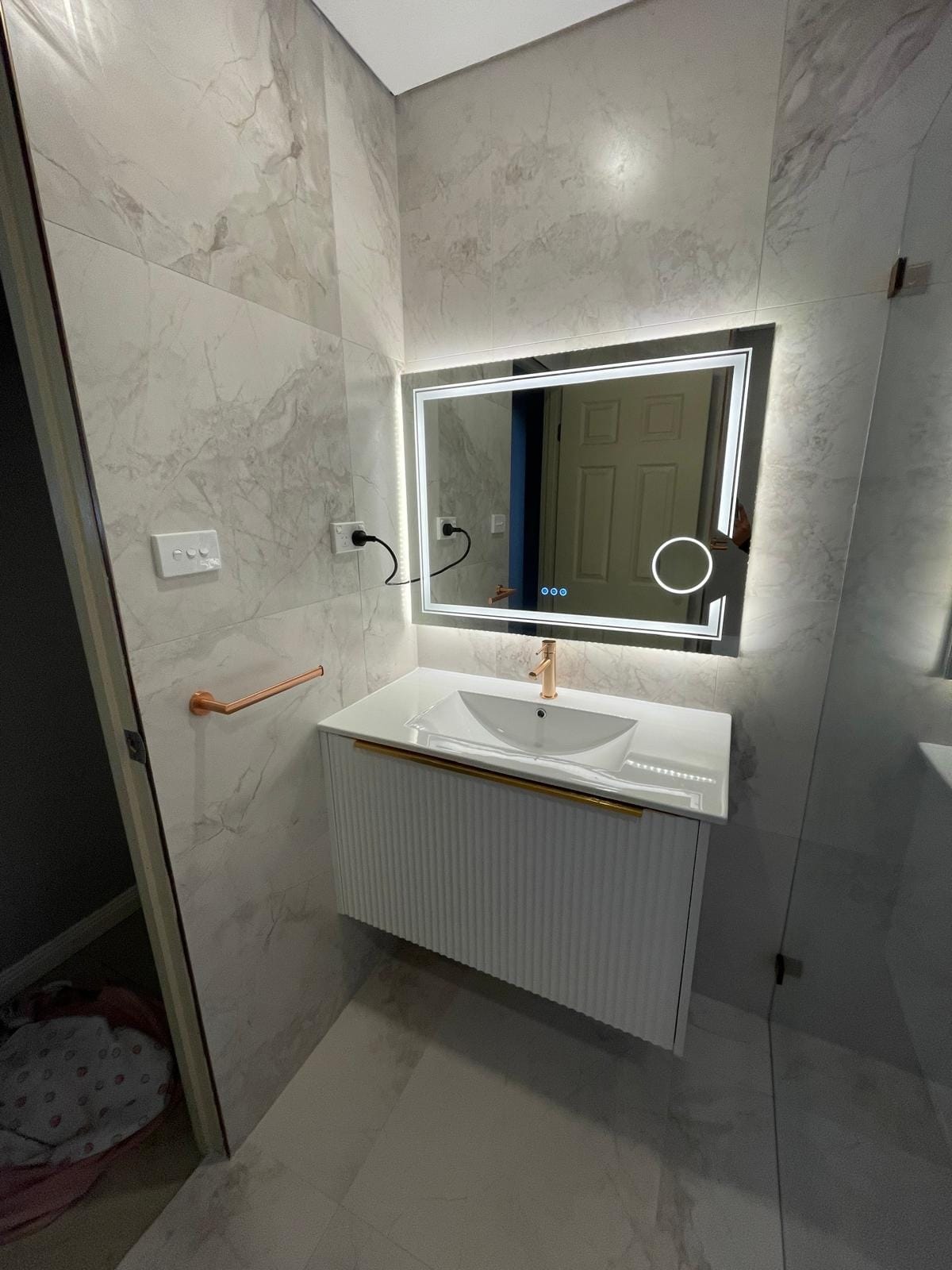 Close view of the fluted vanity: single brass mixer on a moulded basin, backlit mirror with a magnifying inset, and a brass towel rail on the marble-look wall.