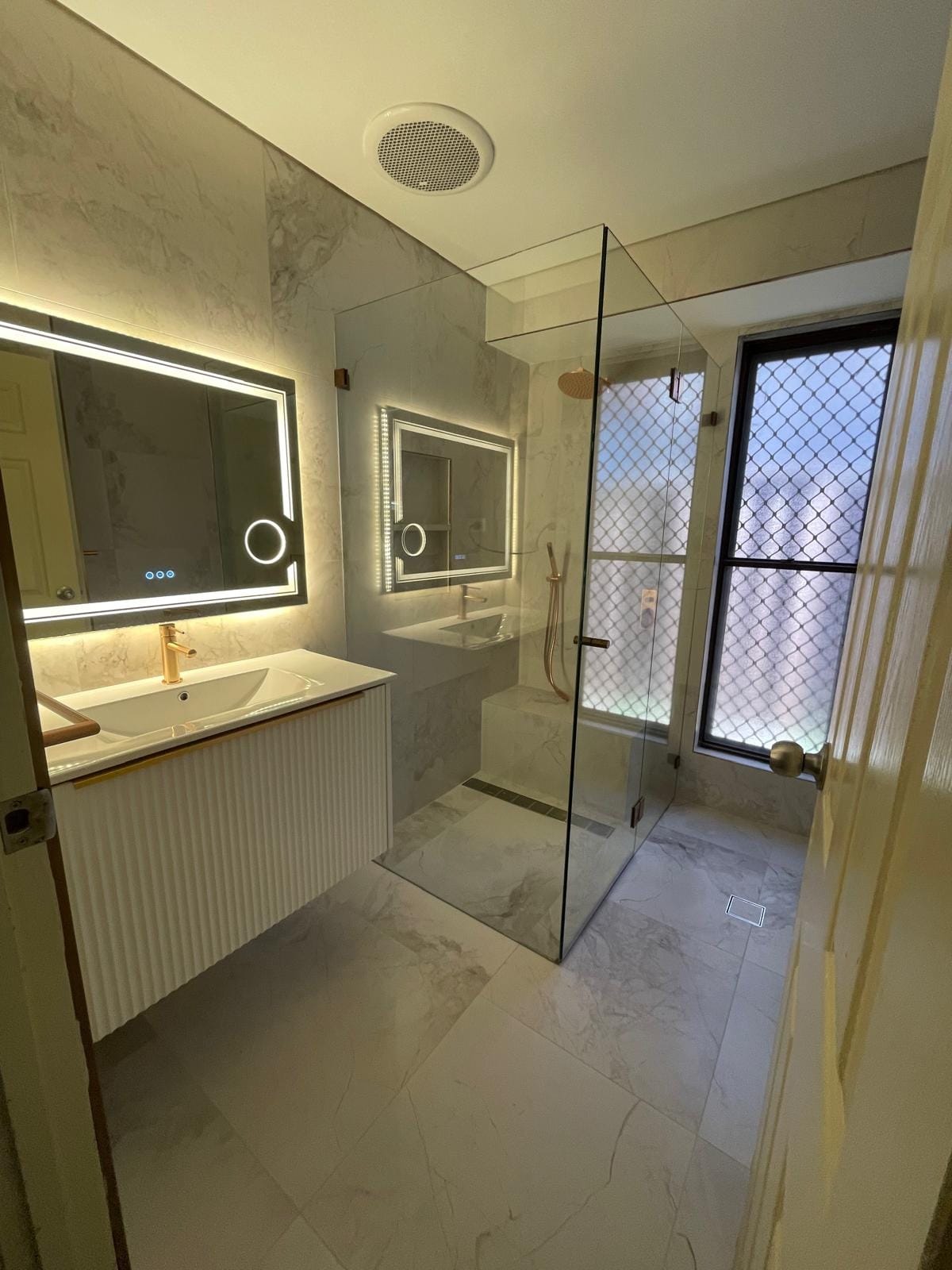 Wide view from the doorway: fluted wall-hung vanity with brass tapware under a backlit mirror, frameless shower beyond and marble-look porcelain throughout.