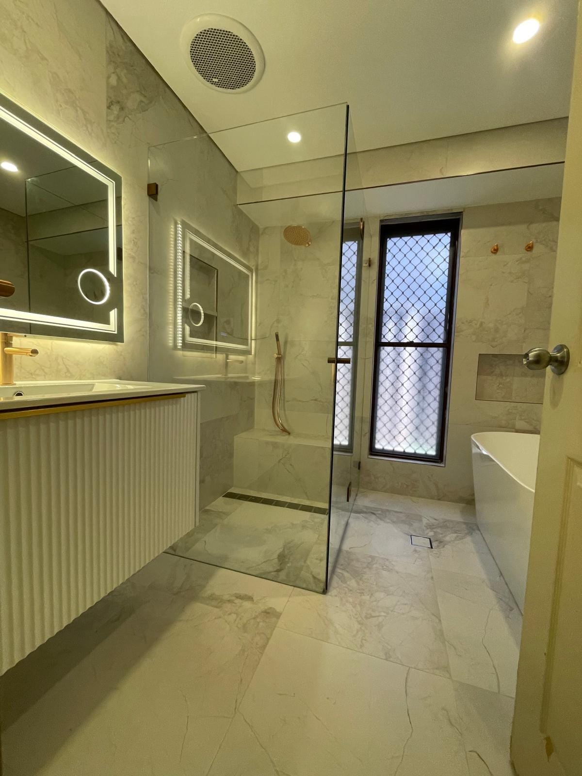 A completed bathroom renovation: marble-look porcelain floor to ceiling, fluted vanity with brass tapware, walk-in shower and a freestanding bath