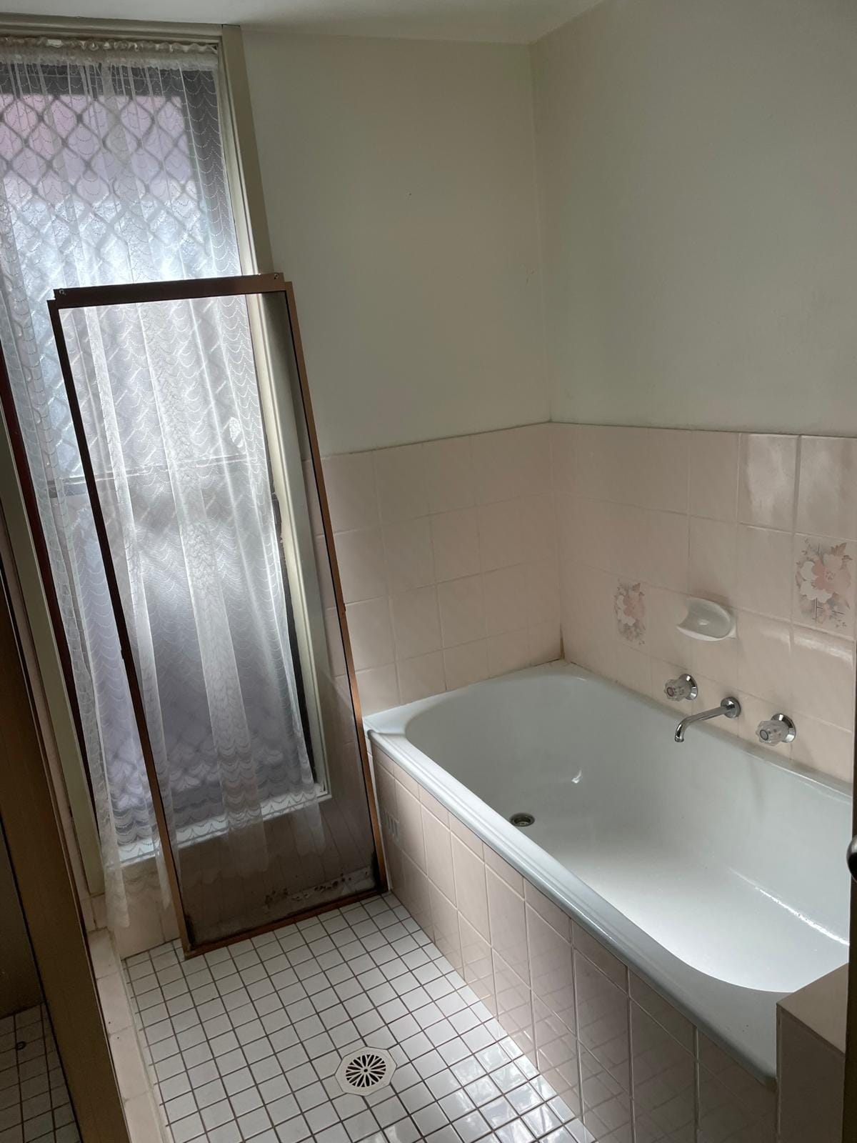 The original bath with chrome wall taps in a tiled surround, pink-cream wall tiles with floral inserts, and a lace-curtained window behind a bronze screen.