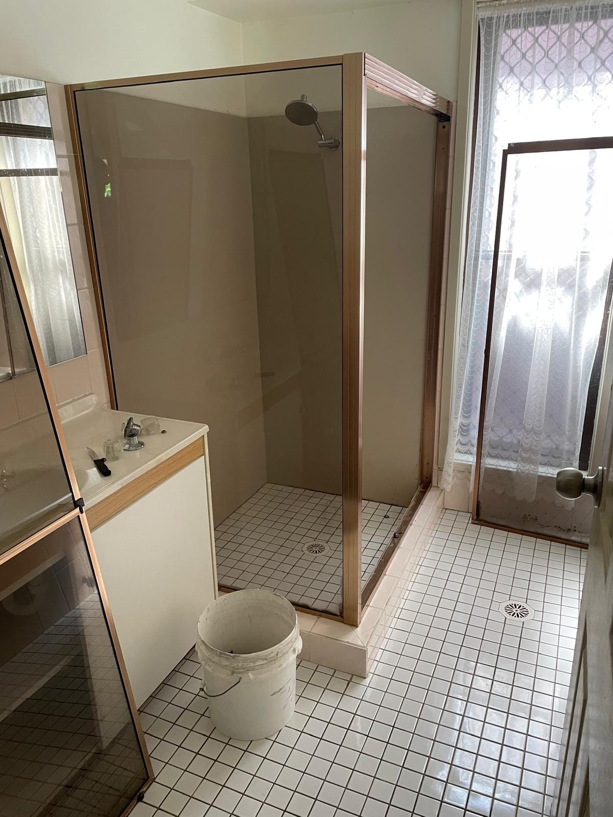 The room as found: a bronze-framed shower screen over small white mosaic floor tiles, laminate vanity and cream wall tiles.