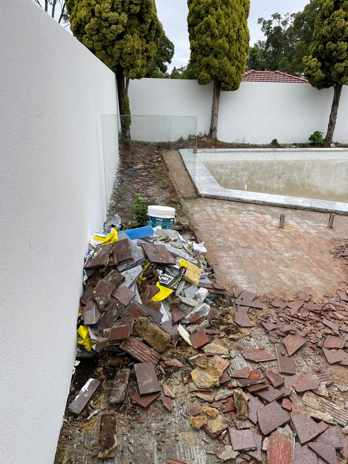 Broken pavers and bedding stacked against the rendered wall for removal, the herringbone bed exposed across the rest of the surround.