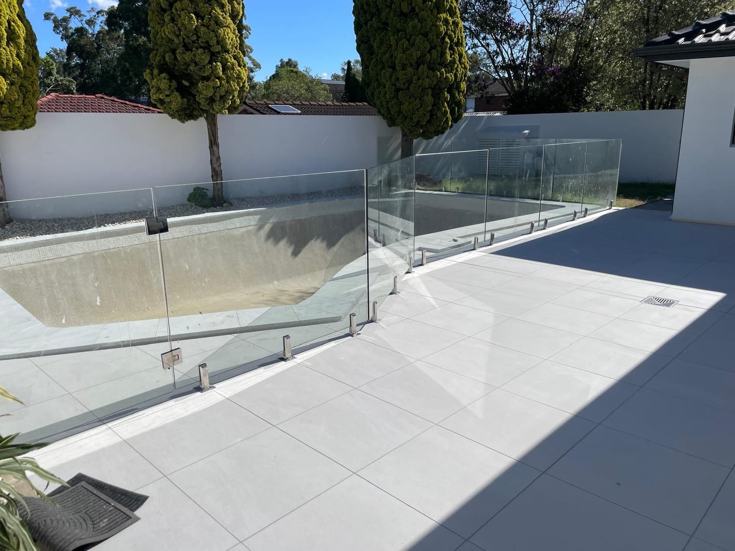 The completed poolside terrace in pale large-format porcelain, frameless glass fence following the water line, rendered boundary wall and conifers behind.