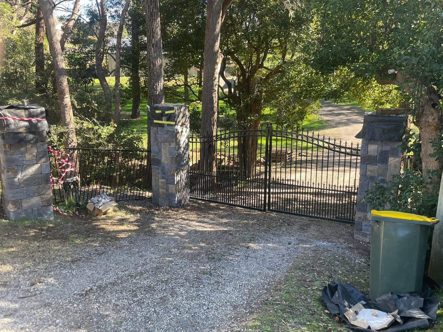 Driveway entry with three piers clad in charcoal stacked stone, wrought iron gates hung between them and bushland either side of the gravel drive.